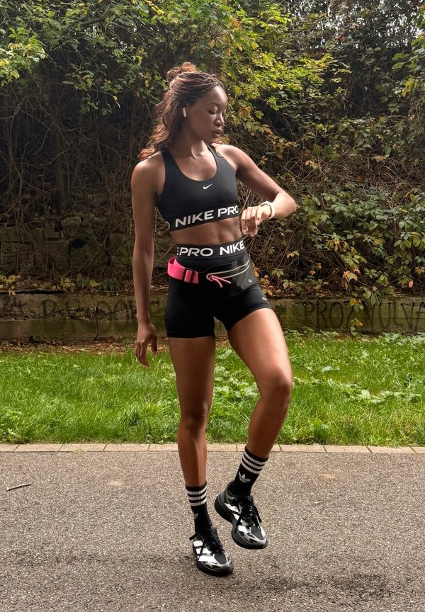Жена с черен спортен екип Nike Pro поглежда китката си на открито край тревиста, залесена местност.