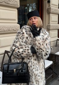 Hvit faux-pelsjakke med leopardmønster, svarte lærhansker og en teksturert svart håndveske. Jakken har et voluminøst design og bred krage.
