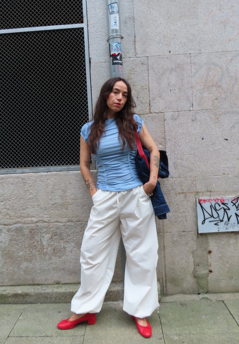 Femme aux longs cheveux debout contre un mur de pierre, portant un haut bleu, un large pantalon blanc et des chaussures rouges, tenant une veste.
