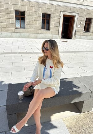 Suéter de punto blanco con acentos de punto coloridos, pantalones cortos claros, sandalias blancas de tacón bloque y gafas de sol. Colocado sobre un banco de piedra al aire libre.