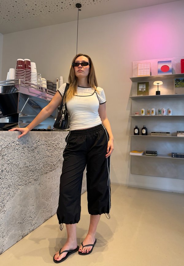 Vrouw draagt een zonnebril, wit t-shirt, zwarte broek en slippers, staat bij een koffietoonbank in een modern café-interieur.