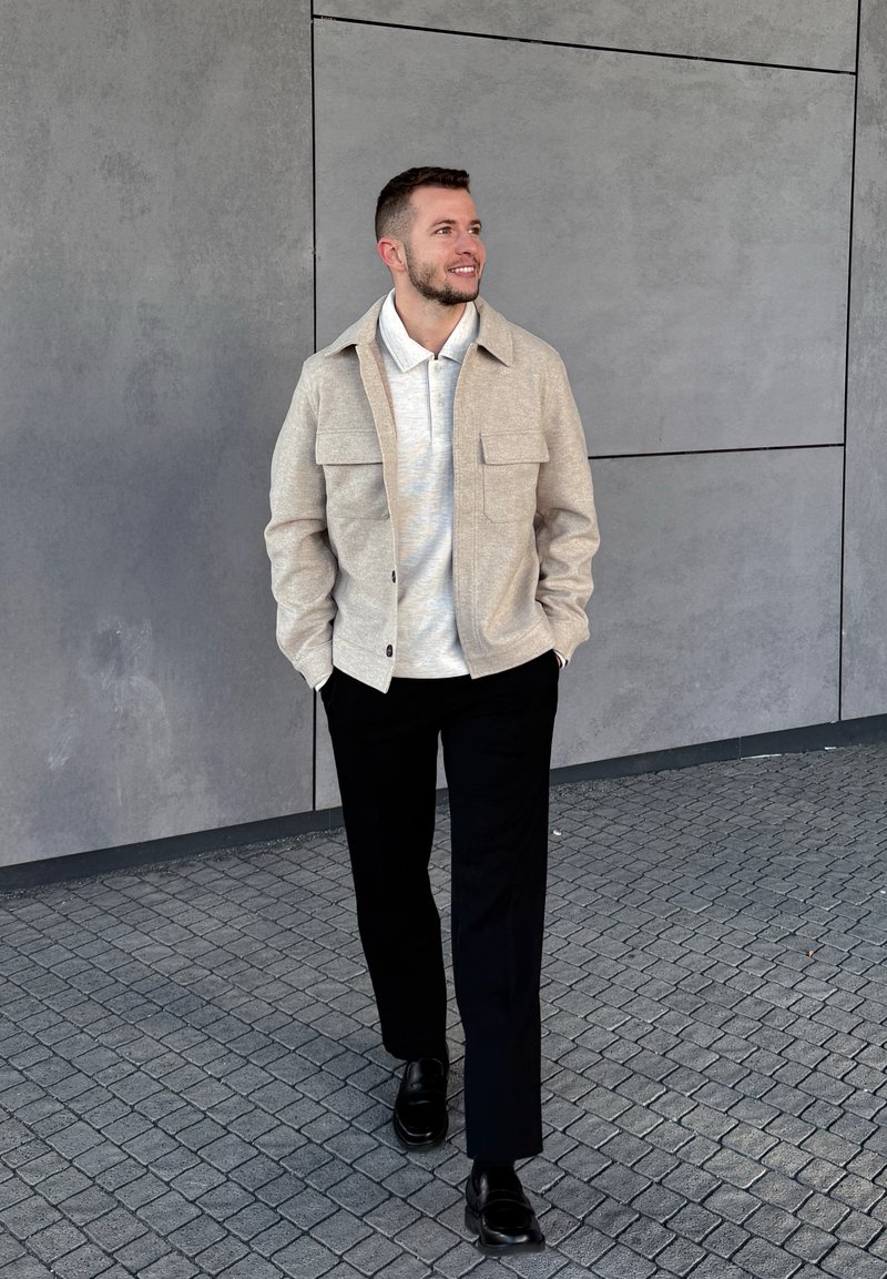 Man walking on tiled pavement wearing beige jacket, white polo shirt, black trousers, and black shoes, against a grey tiled wall.