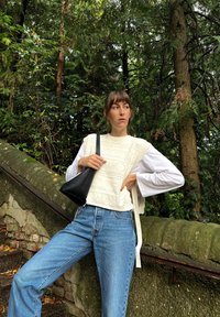 Pulôver branco texturizado sobre uma camisa de manga longa, combinado com jeans azul claro e uma bolsa preta. Fundo verde natural.