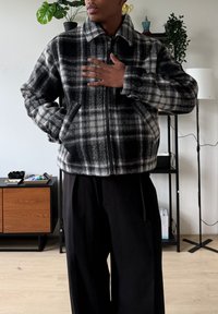 Veste à carreaux noir et blanc avec fermeture éclair, col et deux poches latérales ; en tissu doux et texturé. Portée avec un pantalon noir ample.