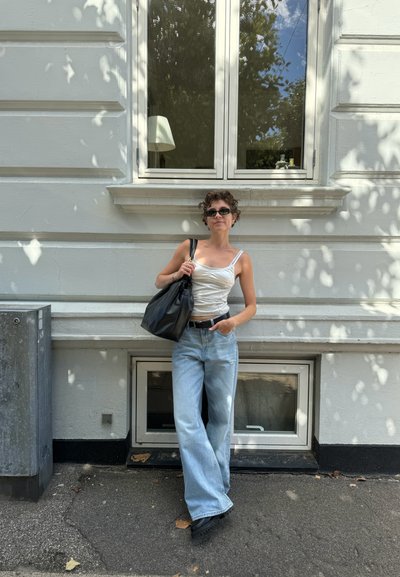 La modelo lleva una blusa blanca de tirantes finos, jeans de pierna ancha de color azul claro y sostiene una bolsa tote negra. El fondo presenta una ventana y una pared gris.