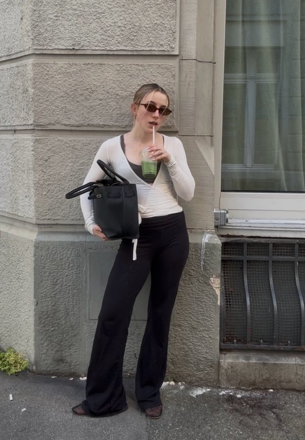 Mladá žena so slnečnými okuliarmi drží čiernu tašku a pije zelené smoothie, keď sa opiera o textúrovanú kamennú stenu pri okne.