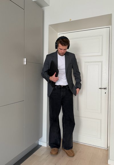 Hombre con auriculares negros, blazer gris, camisa blanca, pantalones anchos negros, zapatos marrones, sosteniendo una funda negra para portátil, de pie junto a una puerta blanca.