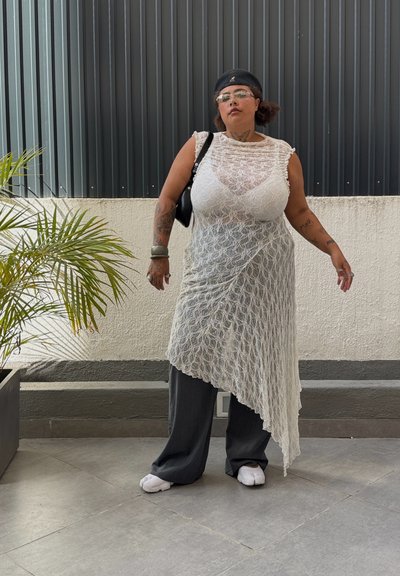 Túnica de encaje en color blanco, corte asimétrico, superpuesta a pantalones anchos grises. Los accesorios incluyen gafas, un bolso negro y joyas.