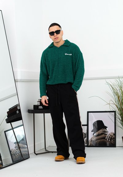 Sudadera verde y borrosa con capucha y costuras laterales, combinada con pantalones deportivos negros y calzado beige; espejos y obras de arte en el fondo.