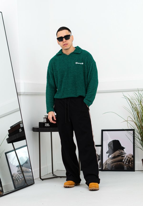 Sweatshirt verde, felpudo, com capuz e costura lateral, combinado com calças de atletismo pretas e calçado bege; espelhos e obras de arte ao fundo.