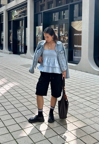 Top peplum azul claro con fruncidos, chaqueta de mezclilla, pantalones cortos de cargo negros y zapatos negros. Bolsa marrón con un encanto, de pie en una calle de baldosas.