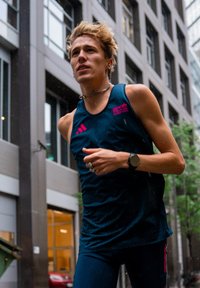 Rövid szőke hajú futó, BMW Berlin Marathon trikót és okosórát visel, ami városi utcán fut esős napon.