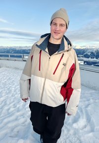 Joven con chaqueta de invierno beige y roja y gorro beige, de pie en una montaña nevada con picos cubiertos de nieve y cielo azul de fondo.