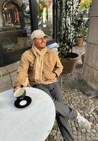 Bež jakna od nubuč kože i šal, sivi hlače, drži crnu šalicu na bijelom okruglom stolu. Vanjski ambijent s zelenilom i pločicama na podu.