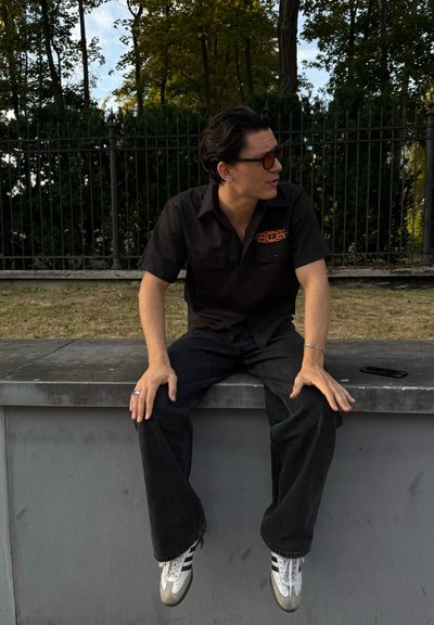 Joven con cabello oscuro y gafas de sol sentado en un borde al aire libre, vistiendo una camiseta negra con logo naranja, jeans negros y zapatillas blancas.