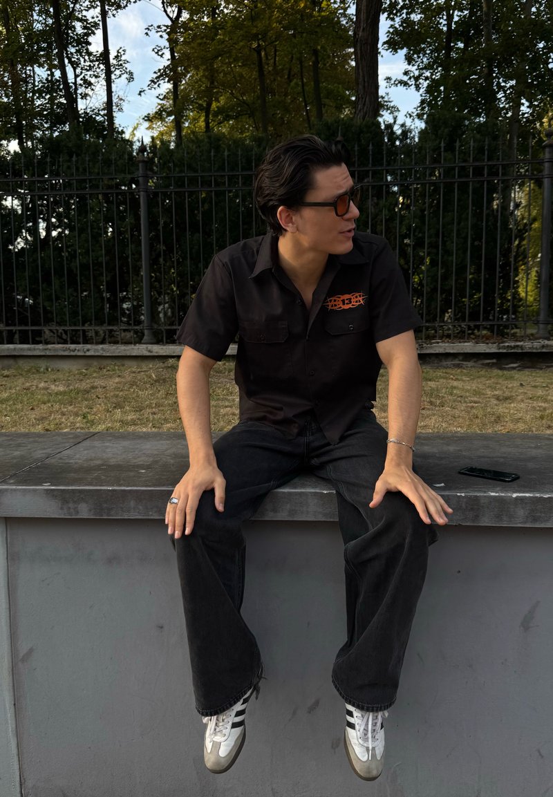 Giovane uomo con capelli scuri e occhiali da sole è seduto su un bordo all'aperto, indossando una maglietta nera con logo arancione, jeans neri e scarpe bianche.