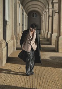 Pantaloni negri cu paiete, blazer bej și un palton lung texturat negru. Modelul pasează pe o suprafață de piatră cu motive sub arcade.