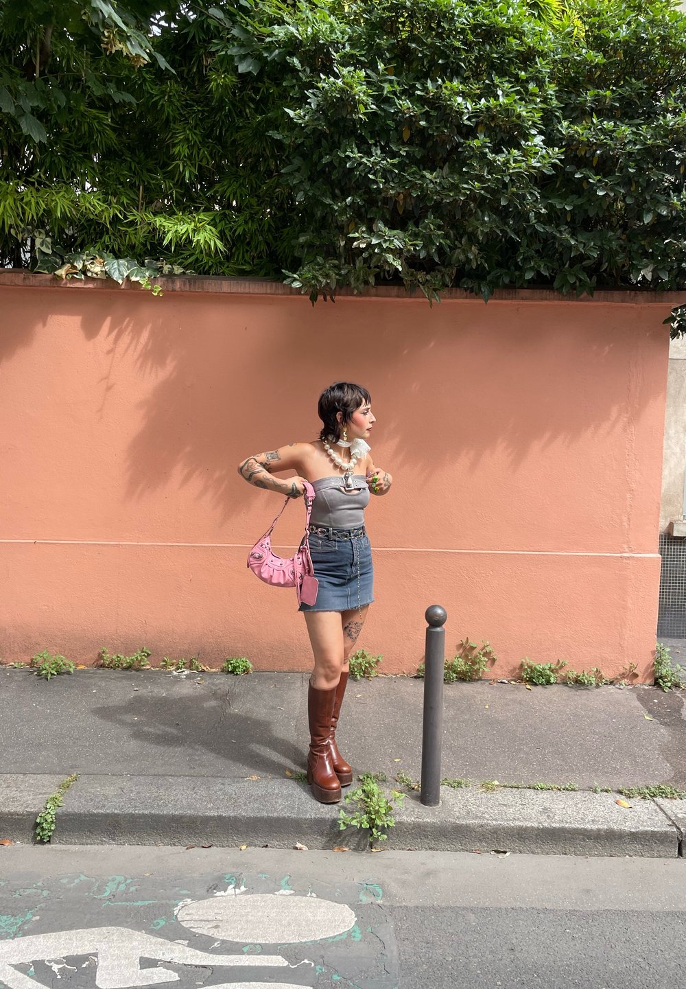 Ψηλές καφέ πλατφόρμες μπότες, τζιν μίνι φούστα, γκρι strapless τοπ, κολάντζα με πέρλες και ροζ τσάντα. Το φόντο έχει έναν ροδάκινο τοίχο με πράσινα φυτά.