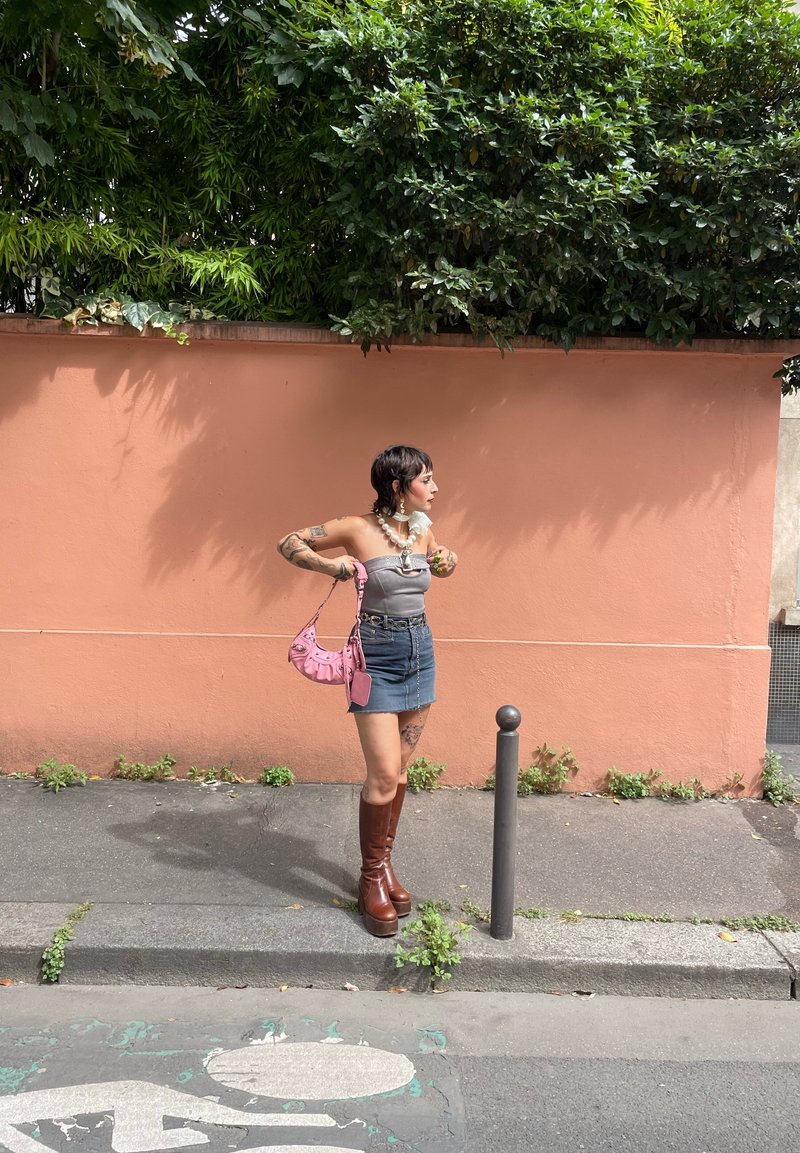 Ψηλές καφέ πλατφόρμες μπότες, τζιν μίνι φούστα, γκρι strapless τοπ, κολάντζα με πέρλες και ροζ τσάντα. Το φόντο έχει έναν ροδάκινο τοίχο με πράσινα φυτά.