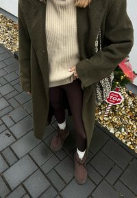 Άτομο που φοράει σκούρο πράσινο παλτό, μπεζ πλεκτό πουλόβερ ριμπ, σκούρα καλσόν, καφέ μπότες, στέκεται σε πλακόστρωτο έδαφος κοντά σε χριστουγεννιάτικη διακόσμηση με πινακίδα "Άγιος Βασίλης στάσου εδώ".