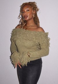 Sweater verde off-the-shoulder com detalhes de franjas, textura canelada e mangas largas. Combinado com jeans pretos ajustados.