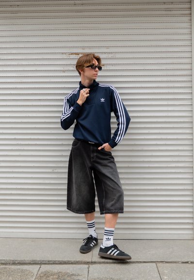 Sudadera navy con tres franjas blancas, pantalones cortos de denim negros, zapatillas negras y calcetines a rayas; look casual de streetwear contra un fondo texturizado.