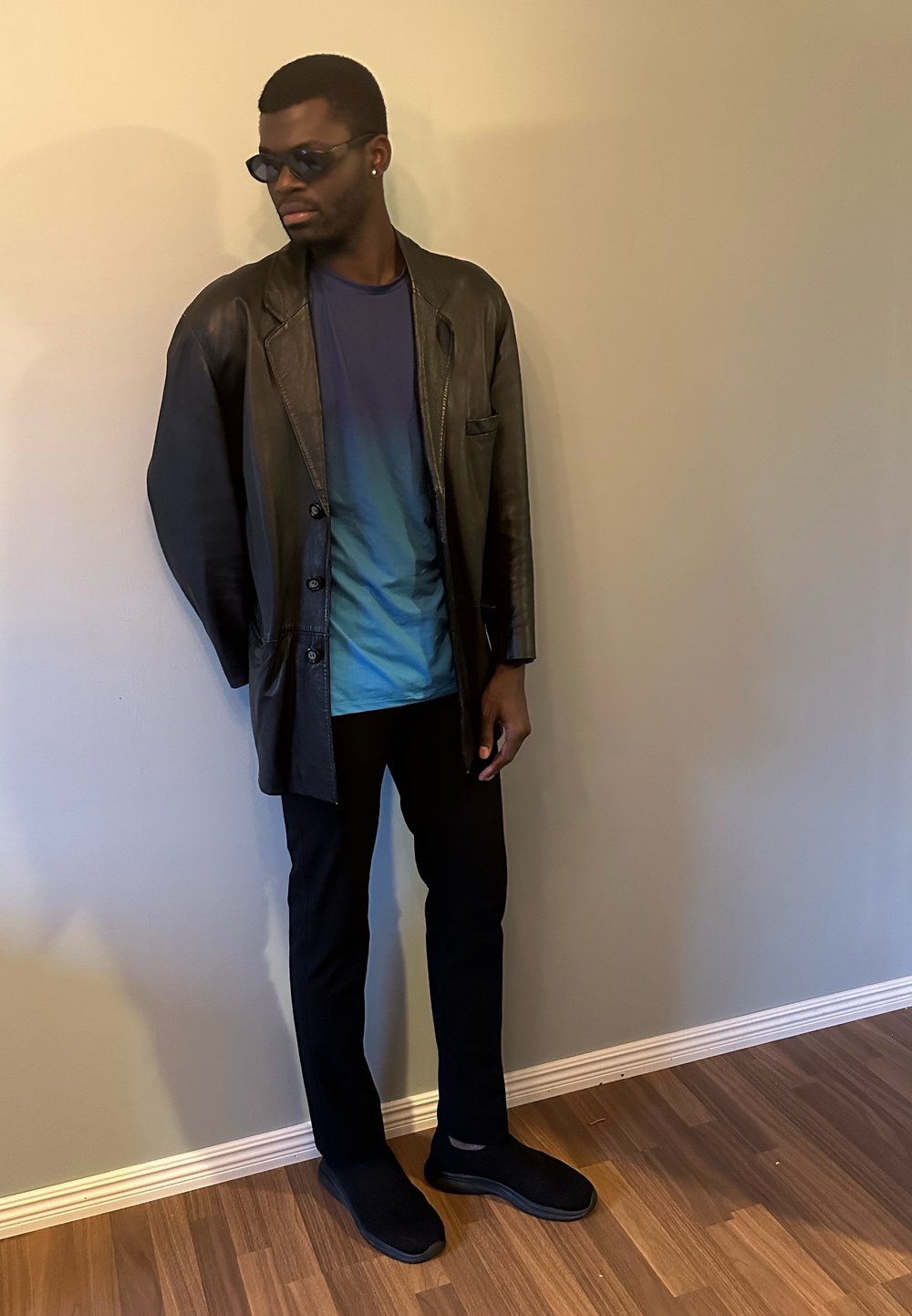 Veste en cuir noire par-dessus une chemise dégradée bleue, pantalon noir et chaussures noires sans lacets. Le mannequin est debout contre un mur de couleur claire.