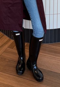 Ben som er iført blå jeans, høye svarte Hunter-gummistøvler og en mørk rubinrød frakk, står på tregulv nær en buet flislagt vegg.