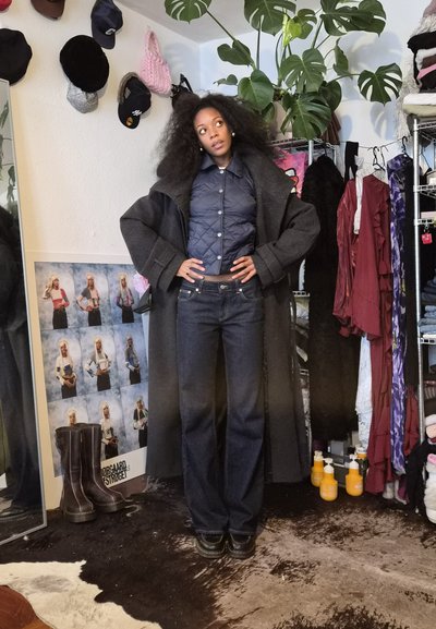 Mujer con cabello rizado y voluminoso, de pie con las manos en las caderas, vestida con un abrigo oscuro grande, chaqueta azul marino acolchada y jeans de pierna ancha en una habitación desordenada.