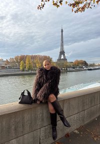 Femme en manteau en fourrure foncée et bottes montant jusqu'aux genoux assise sur une balustrade en pierre près de la Seine, avec la Tour Eiffel et des arbres d'automne en arrière-plan.