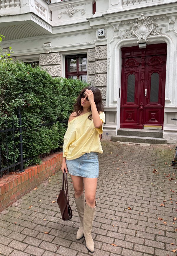 Femme en top jaune épaules dénudées, jupe en jean et bottes beige, debout sur le trottoir tenant un sac marron près d'une porte rouge et d'une haie verte.