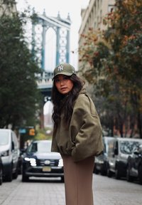 Veste oversize en daim vert olive associée à une jupe beige ajustée. Porte une casquette de baseball verte avec un logo blanc. Décor urbain en arrière-plan.