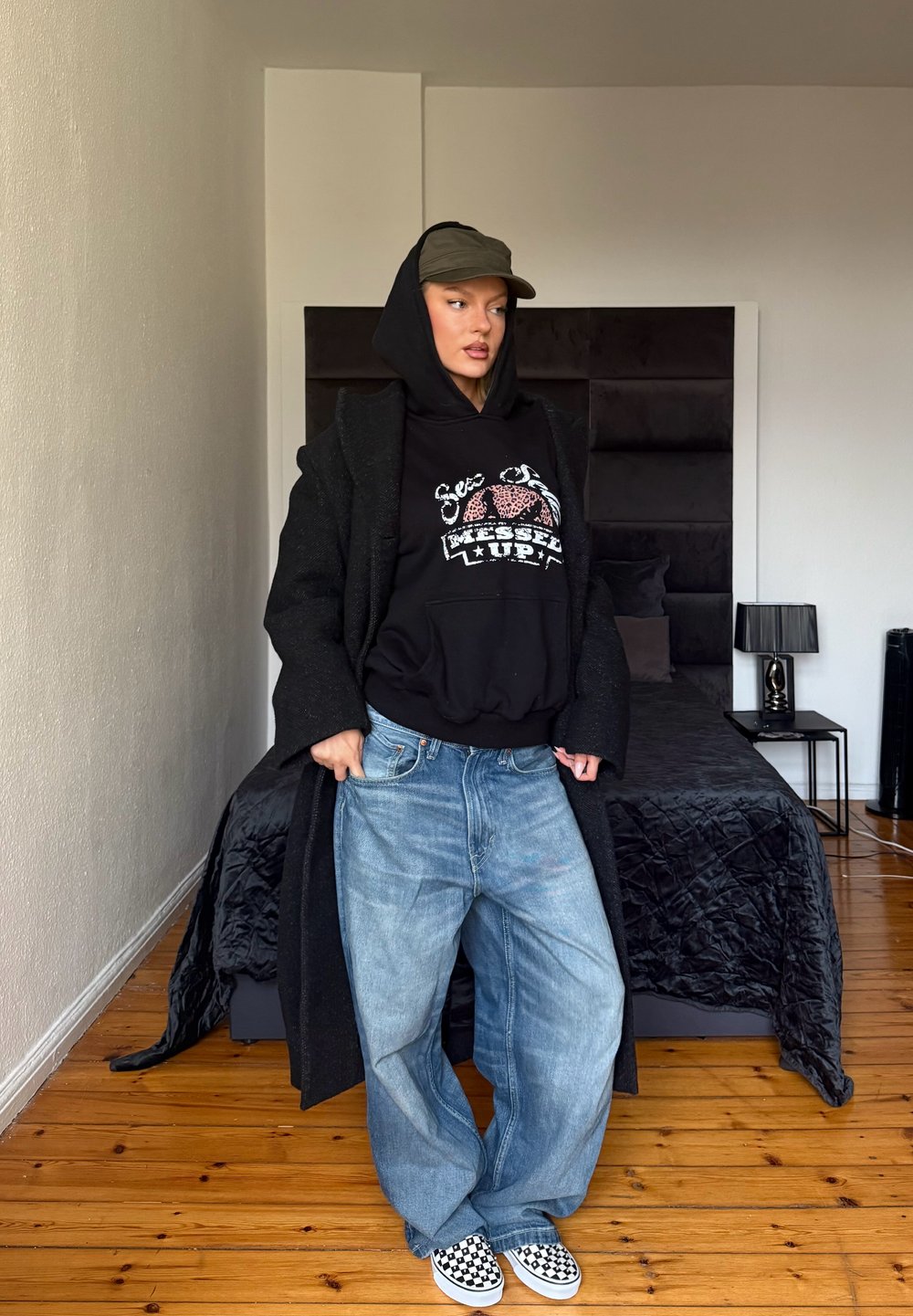 Sweat à capuche noir surdimensionné avec texte graphique, jeans larges bleu clair et chaussures slip-on à carreaux. Manteau en laine et décor moderne de chambre à coucher.