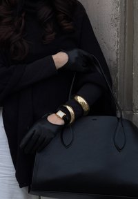 Sac à main en cuir noir aux bords arrondis, tenu par une main gantée en cuir noir. Des bracelets en or et une montre au ton doré complètent le look.