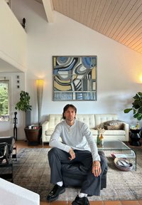 Jongeman met tatoeages die op een zwarte ottomaan zit in een moderne woonkamer met een witte sofa, glazen salontafel en abstracte wandkunst.