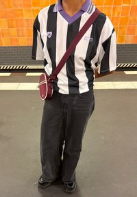 Persona in piedi sulla piattaforma della metropolitana che indossa una maglia a righe bianche e nere con colletto viola e una borsa a tracolla bordeaux.