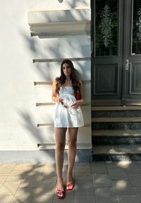 Robe blanche sans manches avec poches latérales, accompagnée de sandales rouges à lanières et d'un sac à main assorti. Debout contre un mur blanc.