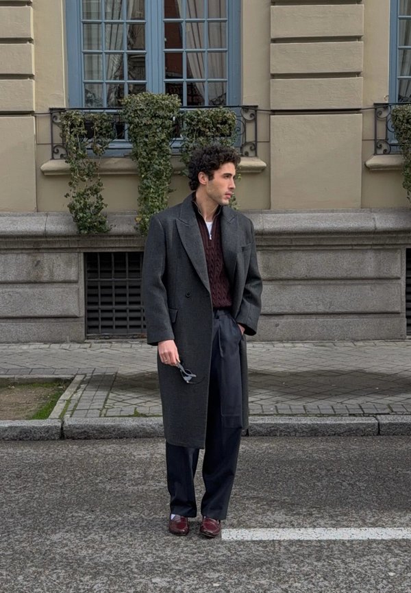 Man with curly hair wearing a dark gray overcoat, burgundy sweater, navy trousers, and burgundy shoes stands on a street holding sunglasses.