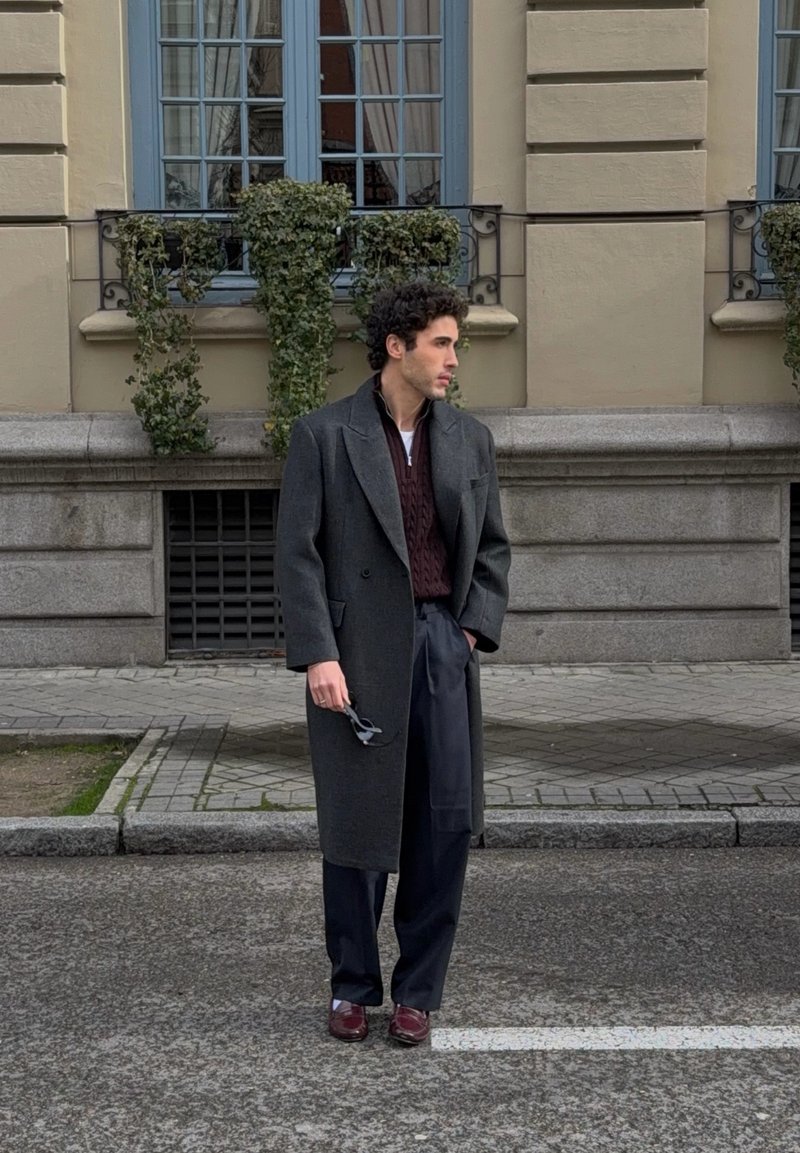 Homme aux cheveux bouclés portant un pardessus gris foncé, un pull bordeaux, un pantalon bleu marine et des chaussures bordeaux, se tenant dans la rue en tenant des lunettes de soleil.