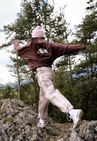Persona che indossa un berretto rosa e una giacca marrone, in equilibrio su un terreno roccioso con le braccia tese, circondata da alberi di pino in un contesto all'aperto.