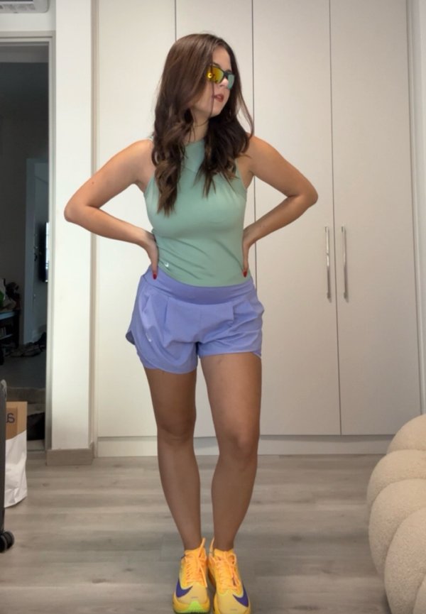 Kvinde står indendørs iført grøn ærmeløs top, lilla shorts, gule sneakers og reflekterende solbriller med hænderne på hofterne.