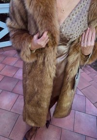 Fuskepelsjakke i brun med en myk tekstur, lagd over en gullfarget satengkjole med broderte blomsterdetaljer, tilbehørt med en sølvfarget clutch.