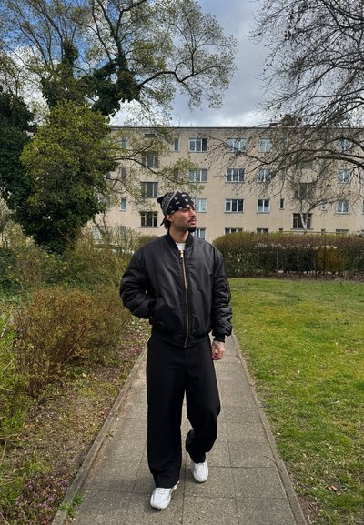 Chaqueta bomber negra, pantalones negros holgados, zapatillas blancas y un pañuelo negro. Entorno al aire libre con vegetación y un edificio de color claro en el fondo.