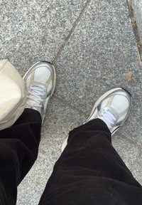 A person wearing white and beige sneakers with black trousers is standing on a grey stone-tiled pavement next to a beige bag.