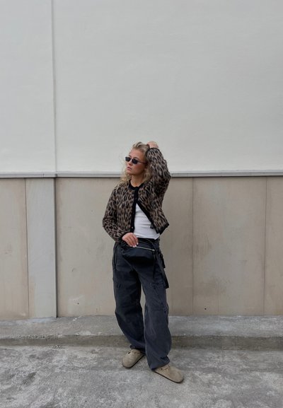 Cardigan de felpa con estampado de leopardo sobre una camiseta negra, combinado con pantalones cargo negros holgados y zapatos de charol marrones, con fondo de una pared neutra.