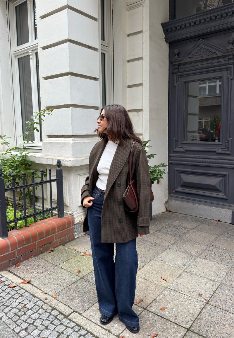 Femme portant des lunettes de soleil, un haut blanc, un blazer marron à carreaux, un jean à jambes larges et des chaussures noires, debout près de l'entrée d'un immeuble sur un trottoir en ville.