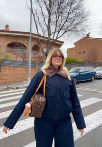 Femme aux cheveux blonds et lunettes, portant un manteau foncé avec un col en fourrure, tenant un sac à main marron à motif, debout sur un passage pour piétons.
