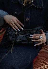 Μαύρη, υφασμάτινη τσάντα ώμου με φερμουάρ και διακοσμητικές καρφίτσες, κρατιέται από χέρια με μακριά, ανοιχτόχρωμα νύχια και πολλούς χρυσούς δακτυλίους.