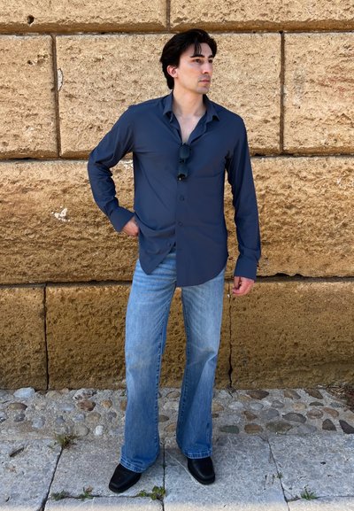 Joven con camisa azul oscuro y gafas de sol en el pecho, vaqueros azul claro y zapatos negros, de pie contra una pared de piedra texturizada.