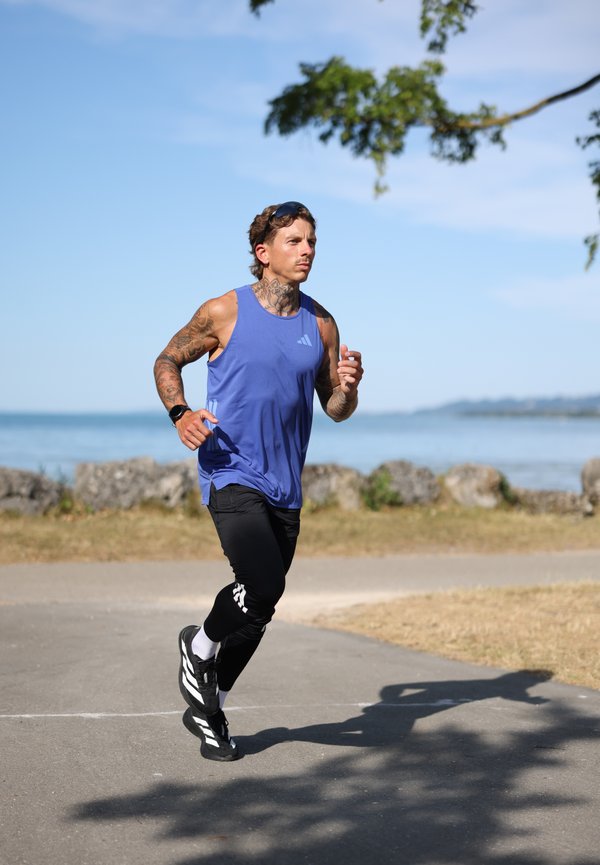 Homme tatoué portant un débardeur bleu et un pantalon noir, faisant son jogging sur un chemin pavé près de l'eau sous un ciel clair.